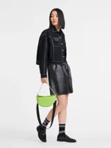 Cartera tipo crossbody de tamaño pequeño, color verde lima, con forma de media luna. Presenta una cadena metálica plateada como asa corta y una correa de cinta negra ajustable y extraíble. Incluye cierre superior y logo de la marca en el frente.
