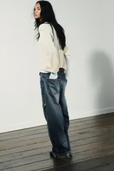 Jeans de mezclilla azul con lavado desgastado y rotos en la pierna derecha. Tienen tiro medio, corte ancho (baggy) y puños doblados en el bajo.