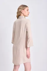 Vestido camisero corto de color beige, con cuello clásico, cierre frontal de botones y detalle de tablas verticales en el frente. Presenta mangas tres cuartos y un corte holgado con ruedo redondeado.