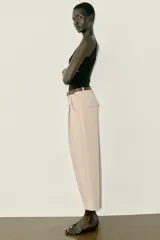 Pantalón tobillero de tiro medio, color beige, con pinzas delanteras y bolsillos frontales. Incluye un cinturón fino de cuero marrón con hebilla metálica dorada y cierre frontal con cremallera, botón interior y gancho.