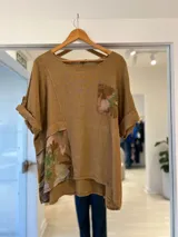 Blusa color camel de lino, con cuello redondo, manga corta con vuelta y detalles de estampado floral en el bolsillo frontal y en el panel inferior.