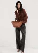 Blusa de color marrón chocolate, confeccionada en tejido plisado con textura. Presenta un diseño de cuello redondo con detalle de lazo, mangas largas amplias con puños acampanados y un corte holgado con volados sutiles en la parte frontal.