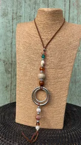 Collar largo tipo gargantilla con tira de cuero marrón, decorado con cuentas de piedras naturales, cuentas plateadas y un dije colgante de dos aros metálicos entrelazados.