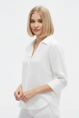 Camisa blanca de algodón, de corte holgado, con cuello camisero, mangas tres cuartos y cierre frontal con botones.