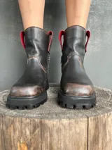 Bota de cuero con efecto desgastado en tonos marrones y negros, con suela track robusta y cierre lateral. Presenta detalles en color rojo en la parte superior de la caña.