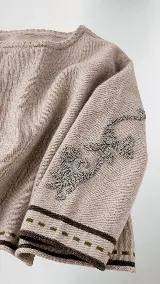 Sweater de lana color beige con calce holgado y cuello redondo. Presenta un bordado negro con diseño de arabescos en una de las mangas y detalles de rayas y puntos en contraste en el ruedo y los puños.