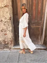 Vestido largo blanco con escote en V profundo y mangas cortas con volantes. Presenta detalles bordados o estampados en hilo dorado en el escote y en los volantes de la falda, con un ajuste fruncido en la cintura.