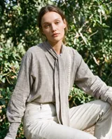 Camisa gris de silueta fluida, confeccionada en tejido texturado, con lazo largo en el cuello, mangas amplias con detalles de puntilla y diseño romántico.