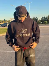 Hoodie de color negro con diseño de estampa frontal que combina una tipografía manuscrita con el texto ITTAI STARS en letras mayúsculas. Presenta un corte holgado, capucha amplia y bolsillo tipo canguro.
