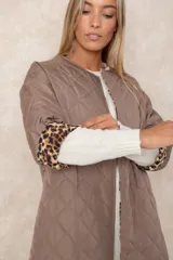 Campera acolchada de corte amplio y largo midi, color beige con diseño capitoneado. Presenta mangas tres cuartos con puños vueltos que revelan un interior estampado con motivo animal print. Diseño sin cuello y cierre frontal oculto.