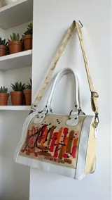 Cartera Diri de cuero natural uruguayo, en tonos marfil y arena, con intervención artística pintada a mano en el frente con pinceladas en rojos, terracotas y dorados. Cuenta con estructura firme, herrajes metálicos en acabado oro viejo, cierres, manijas ergonómicas y correa regulable desmontable.