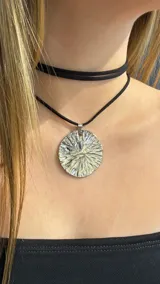 Gargantilla tipo choker de doble vuelta en cordón negro, con un dije circular de metal plateado con textura irregular y acabado brillante.