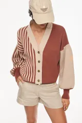 Cardigan tejido bicolor en tonos verde y crudo, con diseño de rayas verticales en la mitad derecha y liso en la izquierda. Presenta cuello en V, cierre frontal con botones y mangas abullonadas con puños ajustados.