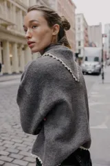 Chaqueta de punto gris oscuro, corte cropped y manga larga, con cuello redondo que incorpora una bandana anudada al frente. Los bordes del cuello, puños y ruedo están terminados con una puntilla de encaje color beige claro. Cierre frontal con botones oscuros.