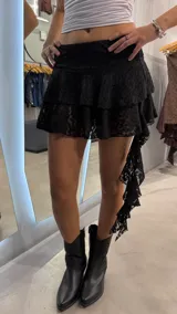 Minifalda de encaje negro con diseño de volados escalonados y detalle de caída lateral asimétrica. Incluye short interno incorporado.