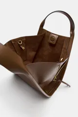Bolso bandolera de formato city color marrón, con diseño estructurado, pespuntes decorativos, doble asa de mano y cremalleras laterales con tiradores colgantes.