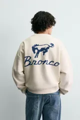 Sudadera de tejido con algodón, color beige claro, de corte relajado (relaxed fit) y cuello redondo. Presenta un parche bordado en el pecho con el logo de Ford y el texto "Genuine Parts" en azul marino.