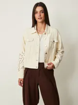 Campera de denim color off white con cuello camisero, bolsillos con solapa en el pecho y cierre frontal con botones metálicos. Presenta detalles de bordado con perforaciones en las mangas.