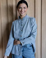 Camisa de denim en tono celeste claro, con cuello alto y detalle de volados a lo largo de la botonadura frontal. Presenta mangas largas con puños abotonados y frunces en los hombros.