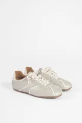 Zapatilla baja de microfibra color gris plateado, con diseño aerodinámico, costuras pespuntadas en contraste blanco, cordones blancos y suela de goma marrón con textura de tapones.