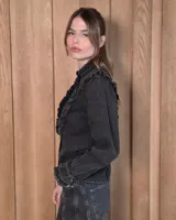 Camisa de denim celeste con cuello alto con volados, botonadura frontal y detalles de volados verticales a lo largo del frente. Cuenta con mangas largas abullonadas y puños con volados.