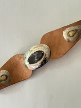 Cinto tipo faja de cuero color marrón, con diseño de piezas de cuero unidas y apliques metálicos plateados grabados con detalles geométricos y una piedra central oscura.