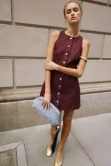 Vestido corto de denim color bordó, sin mangas, con cuello redondo, costuras pespunteadas a contratono y cierre frontal con fila de botones metálicos.