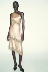 Vestido midi de satén en color beige con escote asimétrico de un solo hombro. Presenta aplicaciones de encaje a tono en el cuerpo y en el bajo, con un diseño de corte asimétrico.