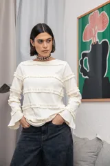 Blusa de color manteca con textura acanalada, cuello redondo y mangas largas con puños acampanados. El diseño presenta múltiples volados horizontales distribuidos a lo largo del cuerpo y las mangas.
