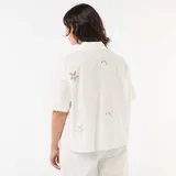 Camisa blanca de algodón de manga corta con cuello camisero, cierre frontal de botones y detalles de bordados calados con formas de estrellas de mar y soles.