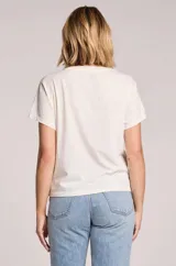 Remera blanca de algodón y poliéster, con cuello redondo y diseño holgado. Cuenta con un estampado frontal de inspiración vintage con la frase "Spritzing the day away", un fondo de tablero a cuadros en tonos celestes y la ilustración de una copa de spritz junto a naranjas y hojas.