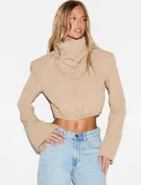 Camisa crop beige de manga larga con cuello alto abotonado, hombreras y puños amplios. Tiene terminación elastizada en la cintura y cierre frontal de botones.