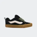 Championes Vans modelo Knu Skool, de color negro con la clásica franja lateral Sidestripe blanca. Presentan diseño robusto de estilo noventero, lengüeta y cuello acolchados, cordones anchos en color verde militar y suela de goma marrón.
