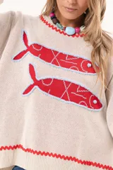 Sweater tejido color crudo con cuello redondo y mangas largas. Presenta dos peces rojos con detalles en azul bordados en el frente. Tiene ribete ondulado rojo en cuello, puños y ruedo.