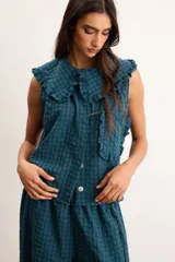 Blusa de algodón con patrón a cuadros vichy en tonos verde oscuro y azul, sin mangas, con cuello amplio decorado con volados, botones delanteros y un bolsillo frontal aplicado con ribete de volados.