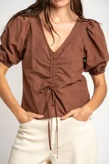 Blusa marrón de algodón con bordados sutiles, escote en V, mangas cortas abullonadas y detalle de fruncido con cordón ajustable en el centro del frente.