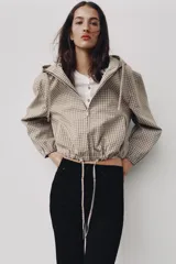 Campera estilo cazadora con capucha ajustable con cordones, cierre frontal de cremallera, mangas largas con puños elásticos, bolsillos delanteros de doble vivo y bajo ajustable con cordones. Presenta un diseño de cuadros en tonos beige y marrón.