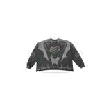 Remera manga larga negra de la colaboración Travis Scott x Fox Racing, con diseño gráfico en tonos grises, marrones y blancos, que incluye el logo de Fox Racing en el pecho, detalles de cadenas y elementos de motocross.
