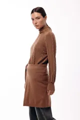 Polera de tejido de punto color marrón, con cuello alto y diseño de trenzas verticales. Presenta mangas largas con puños ajustados y un detalle de panel lateral asimétrico que se anuda en la cintura.