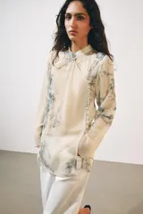 Camisa de cuello redondo y manga larga con diseño de estampado artístico en tonos azules sobre fondo beige. Presenta un cierre frontal asimétrico con botones forrados a tono y una abertura lateral en el escote.