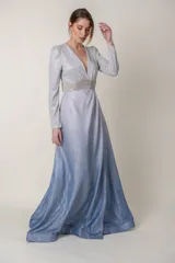 Vestido largo de lurex con acabado brillante en tono lila, con escote en V profundo, mangas largas y cintura marcada con un aplique bordado. La falda presenta un diseño drapeado que nace desde la cintura.