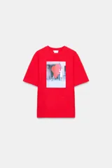 Camiseta de algodón color rojo, corte regular fit, cuello redondo y manga corta. Presenta una estampa fotográfica en el centro con dos personas sosteniendo un gran corazón rojo inflable. Incluye texto con nombre y copyright.