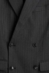 Blazer cruzado de tejido gris oscuro con finas rayas verticales blancas (pinstripe). Presenta solapas de pico, cierre de doble botonadura con botones oscuros, bolsillo superior insertado y dos bolsillos delanteros ribeteados con solapa. Tiene botones decorativos en los puños y abertura trasera.