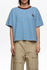 Remera boxy de corte amplio y mangas cortas caídas, confeccionada en tejido de rayas horizontales azules y blancas. Presenta escote redondo con ribete marrón en contraste y un pequeño bordado en el pecho.