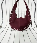 Bolso de hombro tipo hobo color negro, con acabado de gamuza o material similar. Decorado con tachas metálicas redondas plateadas a lo largo del borde inferior y en un dije colgante con forma de corazón.