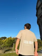 Remera de corte semi oversize en color beige, con estampado gráfico en la espalda que incluye la frase 'BUENA VIBRA' en letras grandes y 'feel the vibes' en tipografía cursiva, además de un pequeño logo bordado en la nuca.