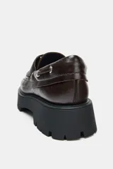 Mocasín de cuero marrón con diseño tipo náutico, presenta ojales metálicos laterales con cordones y suela de plataforma dentada en color negro.
