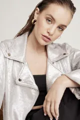 Cazadora estilo biker metalizada en color plateado, con cierre frontal asimétrico, solapas con broches a presión, mangas largas con cierre en los puños y cinturón ajustable en el ruedo.
