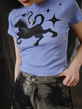 Remera baby tee de calce ajustado y largo corto, color lavanda, con cuello redondo y mangas cortas. Presenta un estampado frontal en serigrafía de ser mitológico y estrellas.
