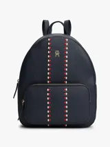 Mochila de diseño minimalista con acabado granulado en color azul marino. Presenta un bolsillo frontal con cierre, correas ajustables, asa superior y el monograma TH metálico en la parte frontal. Se destaca por una cinta decorativa vertical con detalles en rojo y blanco.